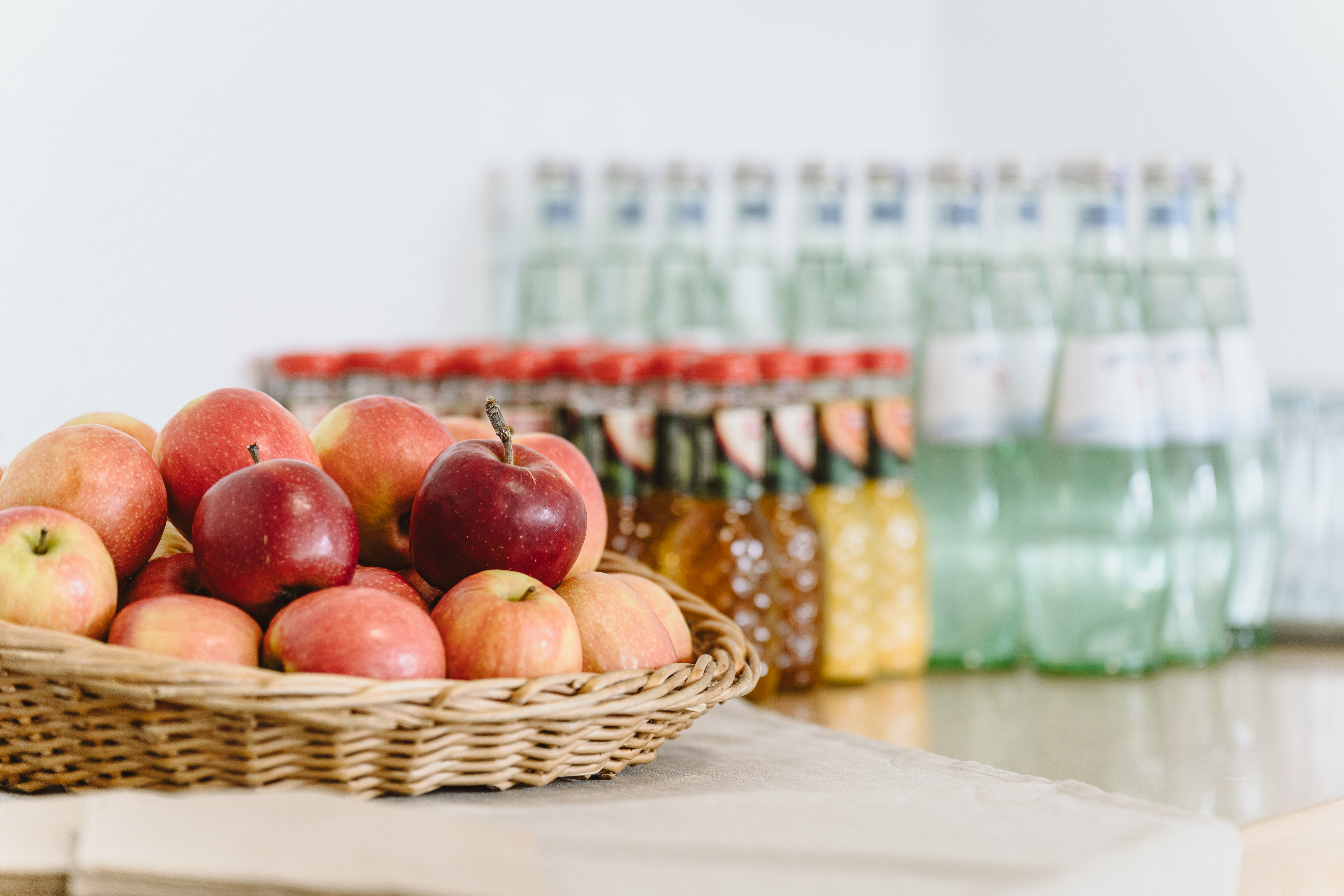 Ein Korb mit frischen, roten Äpfeln, daneben Apfelsaft und Orangensaft in kleinen Flaschen und Mineralwasser.