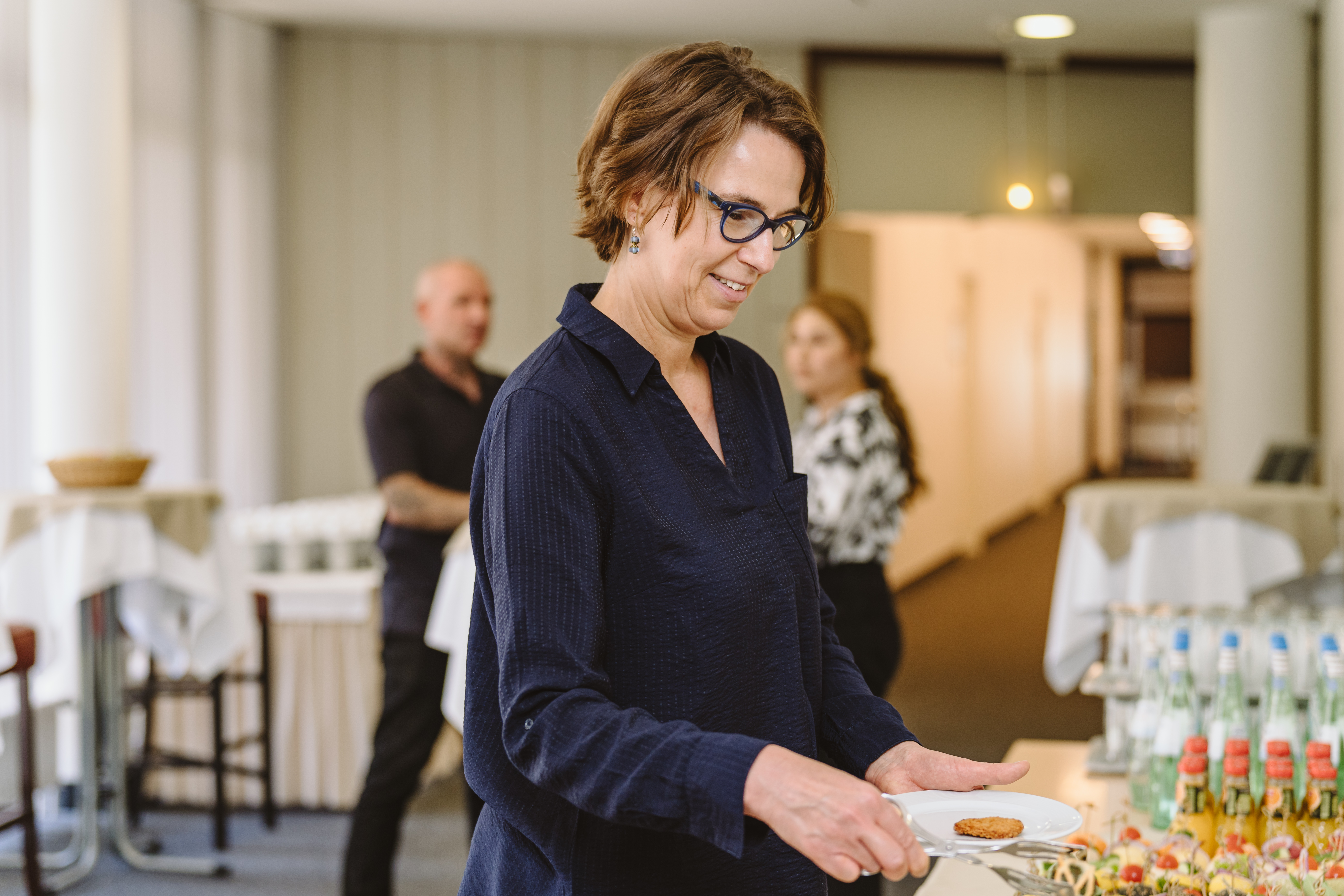 Ein Tagungsgast nimmt sich Snacks vom Buffet. Im Hintergrund unterhalten sich weitere Gäste vor einem Veranstaltungsraum.