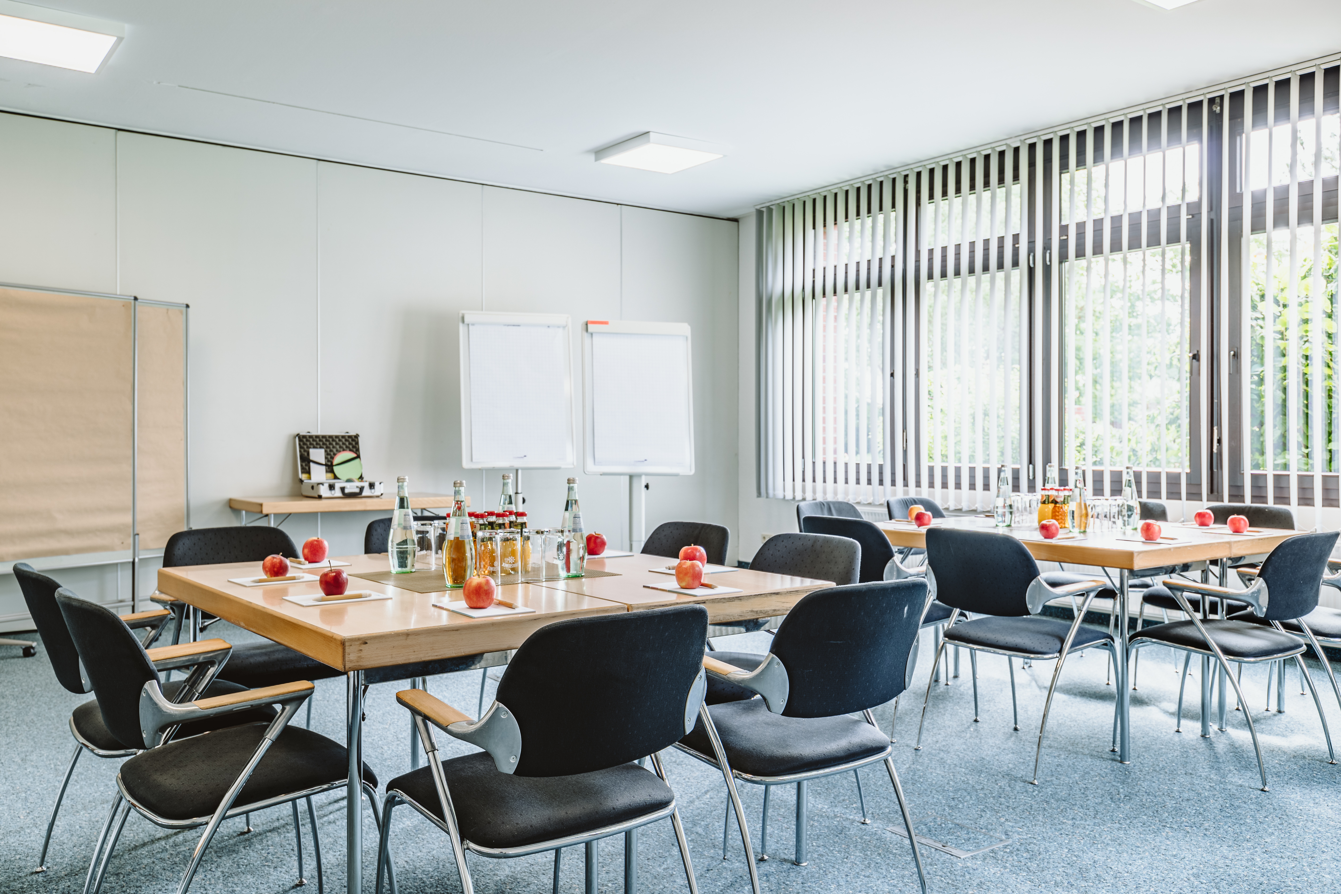 Moderner Tagungsraum mit Flipcharts und Pinnwänden. Die Tische sind in Blockform gestellt und mit Äpfeln und Getränken gedeckt.